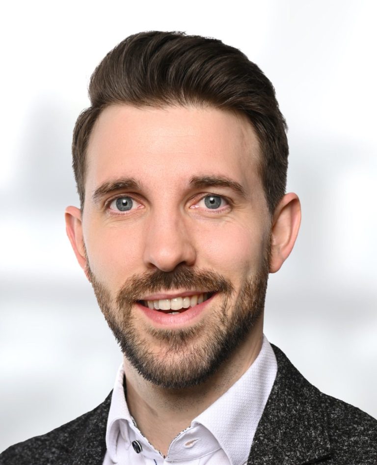 Mediator und Trainer: Matthias Palms Matthias Palms: lächelnder Mann mit kurzer Frisur, Anzug und hellblauer Bluse vor unscharfem Hintergrund.